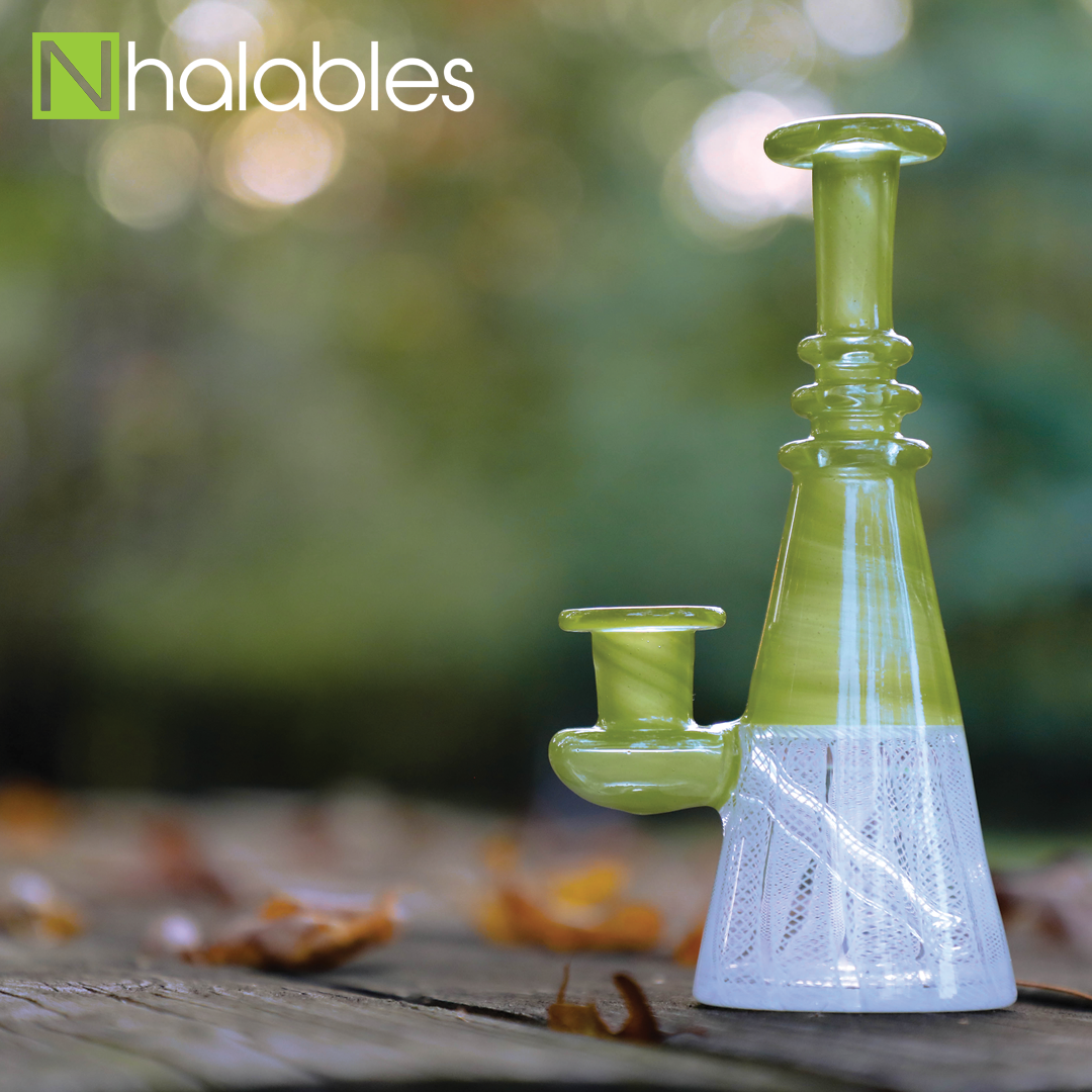 Nhalables Social Posts showing a Jeff Heath Guacamole colored Zanfirico Oil Rig For Concentrate Oils sitting on a picnic table with some fall leaves next to it and blurred trees in the background.