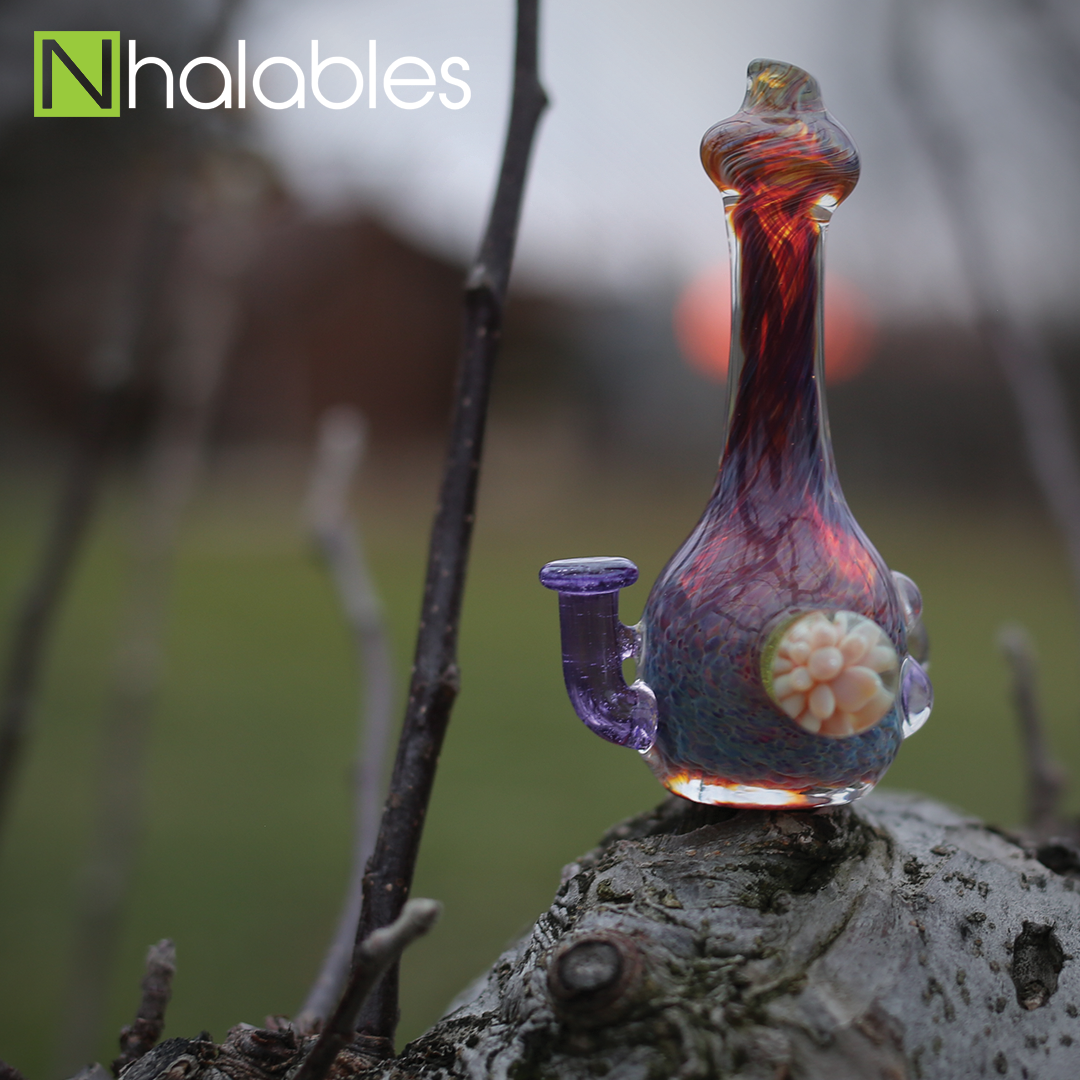 Nhalables Social Post showing a Holden Glass (Josh Holden Brunswick Ohio) fritted chillum sitting on a wooden stump. with grass in the background.