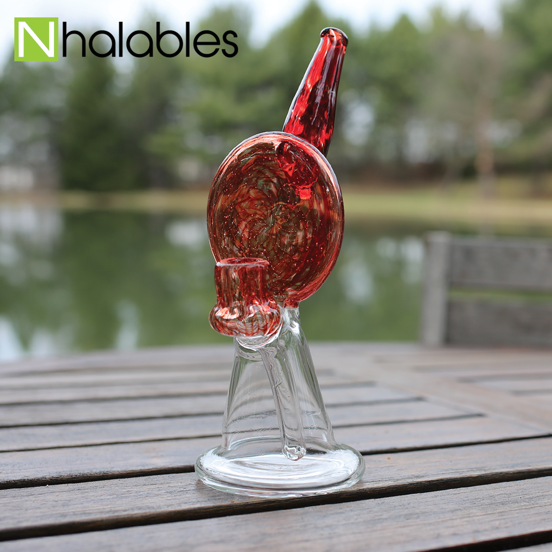 Nhalables Social Post Showing A BTS Glass (Columbia Station Artist, Troy Lawson, Slack Glass) Oil RIg sitting on a wooden table with a pond in the background. 